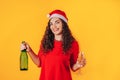 Young beautiful woman on yellow background, on her head she has santa hat. Woman holding bottle and glass in her hands. Christmas Royalty Free Stock Photo