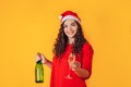 Young beautiful woman on yellow background, on her head she has santa hat. Woman holding bottle and glass in her hands. Christmas Royalty Free Stock Photo