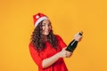 Young beautiful woman on yellow background, on her head she has santa hat. Woman holding bottle in her hands and opening the Royalty Free Stock Photo