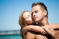 Young beautiful woman embracing and kissing from back her smiling boyfriend with blue sea and sandy beach at background Royalty Free Stock Photo