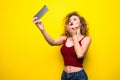 Young beautifulgirl with an curly hairstyle send selfie kiss. Laughing girl take selfie from phone on Yellow background.