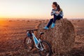 Young bicyclist having rest after a ride in autumn field at sunset. Woman admiring view sitting on haystack Royalty Free Stock Photo
