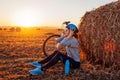 Young bicyclist having rest after a ride in autumn field at sunset. Woman taking off helmet sitting by haystack Royalty Free Stock Photo