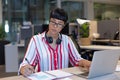 Young biracial businesswoman writing in diary while using laptop at desk in modern workplace Royalty Free Stock Photo