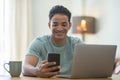 Young black boy using phone and computer connection at home. Happy man reading notification on mobile cellular while work on Royalty Free Stock Photo