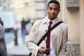 Young black businessman leaning on a wall in the street in London holding phone and smiling to camera, selective focus Royalty Free Stock Photo