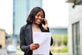 Young Black career woman talking on the phone outside, smiling, holding document Royalty Free Stock Photo