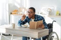 Young black disabled guy in wheelchair eating breakfast and reading book at kitchen. Stay home pastimes