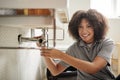 Young black female plumber sitting on the floor fixing a bathroom sink, looking to camera