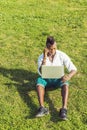 A young black guy with mohawk hair is sitting on a green grasses, working on a laptop computer, making a phone call on his mobile Royalty Free Stock Photo