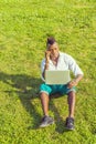 A young black guy with mohawk hair is sitting on a green grasses, working on a laptop computer, making a phone call on his mobile Royalty Free Stock Photo