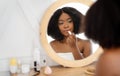 Young black lady applying lipstick in front of mirror at home, putting on decorative makeup. Daily beauty routines Royalty Free Stock Photo