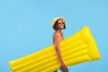 Young black lady in swimwear and straw hat holding yellow inflatable lilo on blue studio background Royalty Free Stock Photo