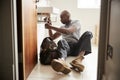 Young black male plumber sitting on the floor fixing a bathroom sink, seen from doorway, full length