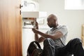 Young black male plumber sitting on the floor fixing a bathroom sink, seen from doorway