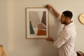 Young black man hanging painting, putting picture frame on wall indoors. Modern home decor and interior design Royalty Free Stock Photo