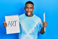 Young black man holding art notebook and painter brushes sticking tongue out happy with funny expression