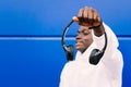 Young black man holding wireless headphones while wearing a white sweatshirt, against a blue wall looking away with confidence Royalty Free Stock Photo