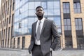 Young black man manager in suit holding hands in trousers pockets, standing against business center