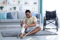 Young black man suffering from pain in plastered broken leg, sitting on floor after falling with crutches, indoors