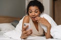 Young black surprised woman using smartphone while lying in bed