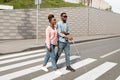 Young black woman assisting visually impaired millennial guy with cane crossing city street. Vision disability concept