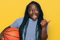 Young black woman with braids holding basketball ball pointing thumb up to the side smiling happy with open mouth Royalty Free Stock Photo