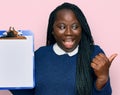 Young black woman with braids holding clipboard with blank space pointing thumb up to the side smiling happy with open mouth Royalty Free Stock Photo