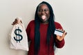 Young black woman with braids holding dollars bag and credit card sticking tongue out happy with funny expression Royalty Free Stock Photo