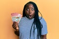 Young black woman with braids holding 100 new zealand dollars banknote scared and amazed with open mouth for surprise, disbelief Royalty Free Stock Photo