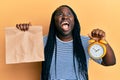 Young black woman with braids holding take away food and alarm clock angry and mad screaming frustrated and furious, shouting with Royalty Free Stock Photo