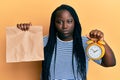 Young black woman with braids holding take away food and alarm clock skeptic and nervous, frowning upset because of problem Royalty Free Stock Photo