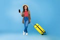 Young black woman with suitcase showing smartphone with blank screen, offering mockup, booking summer vacation online