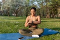 Young black woman using cellphone, listening to music, checking fitness tracker during outdoor yoga training at park Royalty Free Stock Photo