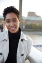 Young black woman wearing a turtleneck sweater and a mackintosh standing on Millennium Bridge, London, looking to camera smiling, 