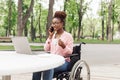 Young black woman in wheelchair working online, using laptop, speaking on smartphone and enjoying coffee at outdoor cafe Royalty Free Stock Photo