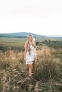 Young blond beautiful hippie girl in white dress and feather hair accessories on summer field and mountains background Royalty Free Stock Photo