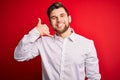 Young blond businessman with beard and blue eyes wearing elegant shirt over red background smiling doing phone gesture with hand Royalty Free Stock Photo