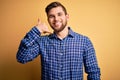 Young blond businessman with beard and blue eyes wearing shirt over yellow background smiling doing phone gesture with hand and Royalty Free Stock Photo