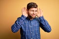 Young blond businessman with beard and blue eyes wearing shirt over yellow background Trying to hear both hands on ear gesture, Royalty Free Stock Photo