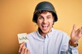 Young blond businessman with curly hair wearing helmet holding paper with war message very happy and excited, winner expression Royalty Free Stock Photo