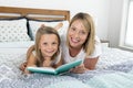 Young blond Caucasian mother lying on bed with her young sweet 7 years old daughter reading book together at home bedroom Royalty Free Stock Photo