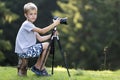 Young blond child boy sitting on tree stump on grassy clearing taking picture with tripod camera