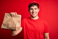 Young blond delivery man with curly hair wearing cap holding paper bag with food with a happy face standing and smiling with a Royalty Free Stock Photo