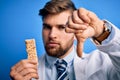 Young blond doctor man with beard and blue eyes wearing coat eating granola bar with angry face, negative sign showing dislike Royalty Free Stock Photo