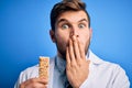 Young blond doctor man with beard and blue eyes wearing coat eating granola bar cover mouth with hand shocked with shame for Royalty Free Stock Photo