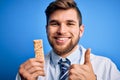 Young blond doctor man with beard and blue eyes wearing coat eating granola bar happy with big smile doing ok sign, thumb up with Royalty Free Stock Photo