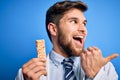 Young blond doctor man with beard and blue eyes wearing coat eating granola bar pointing and showing with thumb up to the side Royalty Free Stock Photo