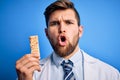 Young blond doctor man with beard and blue eyes wearing coat eating granola bar scared in shock with a surprise face, afraid and Royalty Free Stock Photo