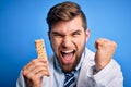 Young blond doctor man with beard and blue eyes wearing coat eating granola bar screaming proud and celebrating victory and Royalty Free Stock Photo